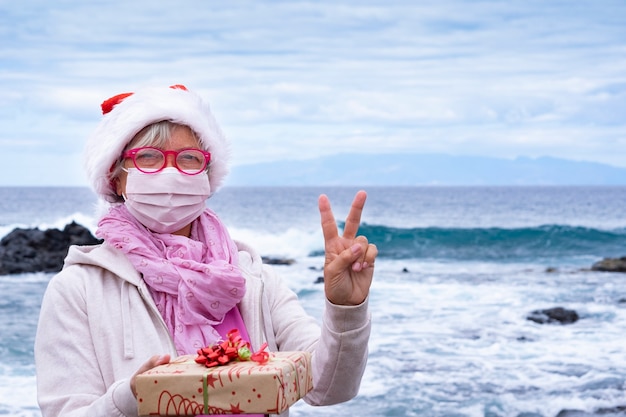 Senior vrouw in een kerstmuts met een beschermend masker op het strand met kerstcadeau