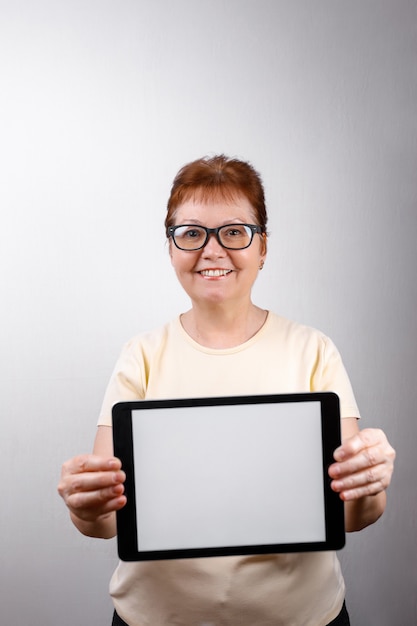 Senior vrouw in glazen toont een tablet op wit in een licht t-shirt