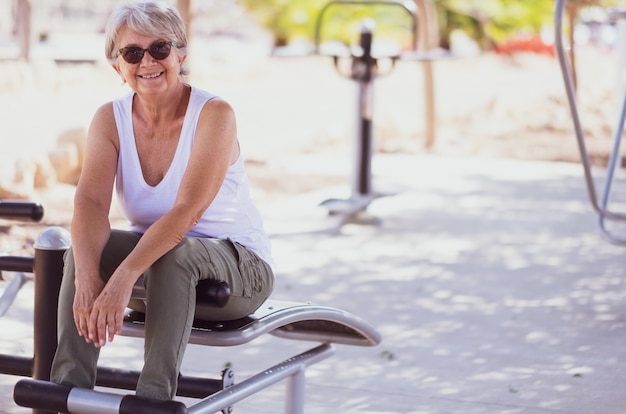 Senior vrouw in gymnastiek activiteit in openbaar park, zittend op een buikbankje kijkt glimlachend naar de camera