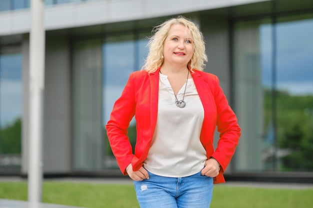 Senior vrouw in rode jas blouse en spijkerbroek in de buurt van het moderne gebouw