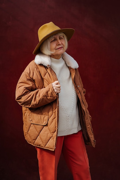 Senior vrouw in trendy bovenkleding en hoed die wegkijkt terwijl ze tegen de wijnmuur staat.
