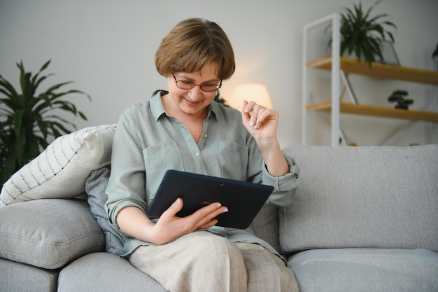 Senior vrouw kijkt en lacht om haar digitale tablet op de bank