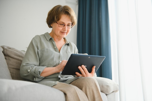 Senior vrouw kijkt en lacht om haar digitale tablet op de bank