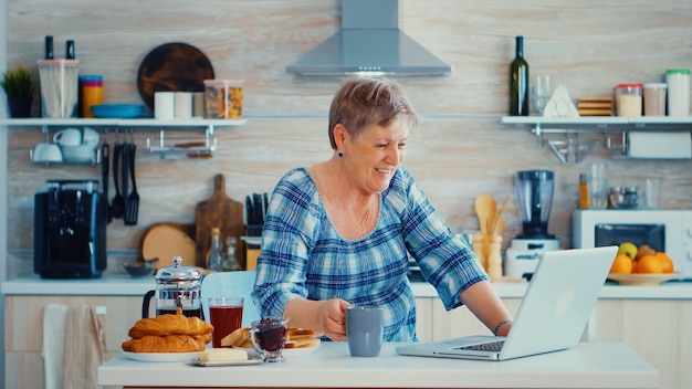 Senior vrouw koffie drinken en zwaaien tijdens een videoconferentie met familie in de keuken tijdens het ontbijt. Oudere persoon die internet online chattechnologie gebruikt, videowebcam die een videogesprek voert c