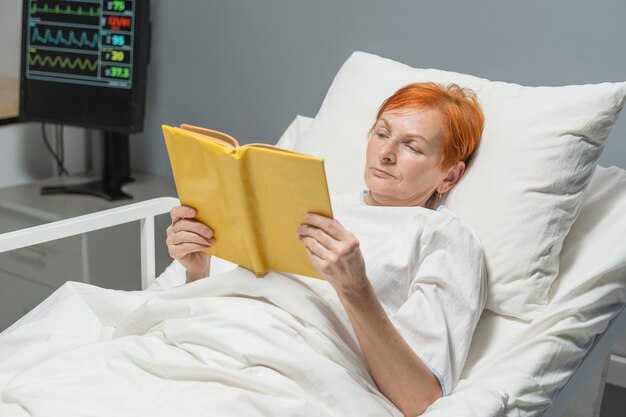 Senior vrouw liggend op bed in ziekenhuisafdeling en het lezen van een boek, ze herstellende na ziekte