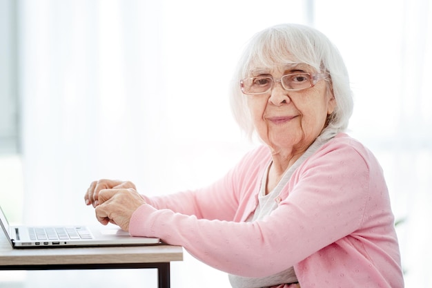 Senior vrouw met behulp van laptop thuis en kijken naar de camera. Bejaarde met moderne technologieën portret