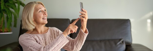 Senior vrouw met behulp van mobiele telefoon zittend op de bank. Oudere vrouw die op de bank zit en een telefoonbericht sms't.