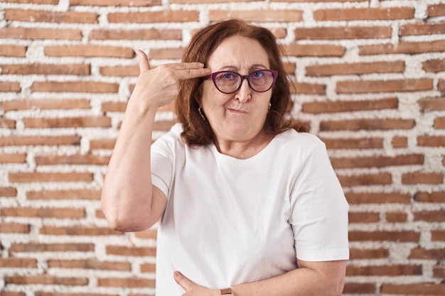 Senior vrouw met bril staande over bakstenen muur schieten en zelfmoord plegen wijzende hand en vingers naar het hoofd als pistool zelfmoord gebaar