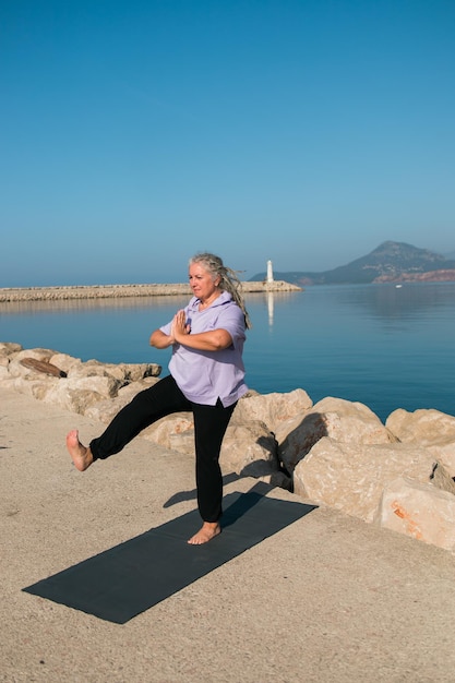 Senior vrouw met dreadlocks in stretch positie aan zee in de ochtend Bejaarde vrouw doet yoga in de buurt van het strand