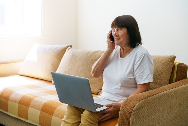 Senior vrouw met laptopcomputer zittend op de bank praten met iemand aan de telefoon nieuwe technologieën zijn ...