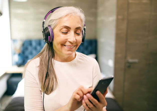 Senior vrouw met wit haar in koptelefoon zet muziek op de smartphone