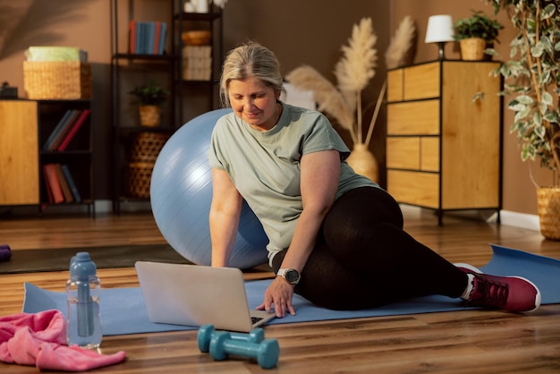 Senior vrouw oma zittend op yoga mat rustend surfen op internet op zoek naar yoga oefening