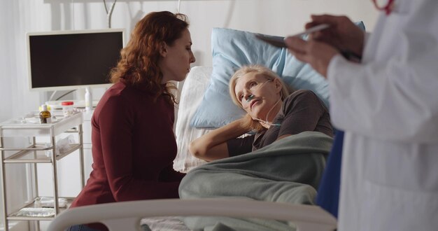 Foto senior vrouw patiënt liggend in ziekenhuisbed met volwassen dochter zitten in de buurt en luisteren naar arts