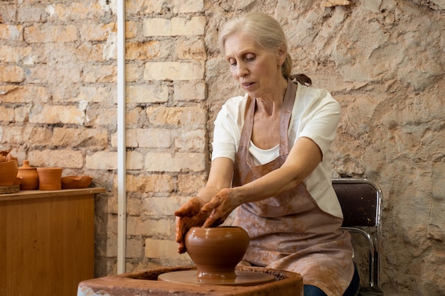 Senior vrouw pottenbakker beeldhouwt een aarden pot de beeldhouwer werkt met klei op een pottenbakkersschijf en aan een tafel