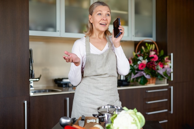 Senior vrouw praten over de telefoon in de keuken en bereiden van voedsel