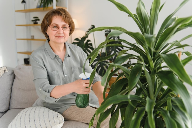 Senior vrouw spuit planten in bloempotten Gelukkige vrouw zorgt voor kamerplant Rijpe vrouw zorgt voor planten bij haar thuis en sproeit een plant met zuiver water uit een spuitfles