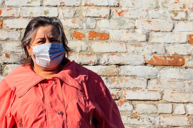 Senior vrouw staat met een medisch masker op haar gezicht, dat beschermt tegen coronavirus en andere virussen en ziekten op een achtergrond van oude bakstenen muur. coronapandemie