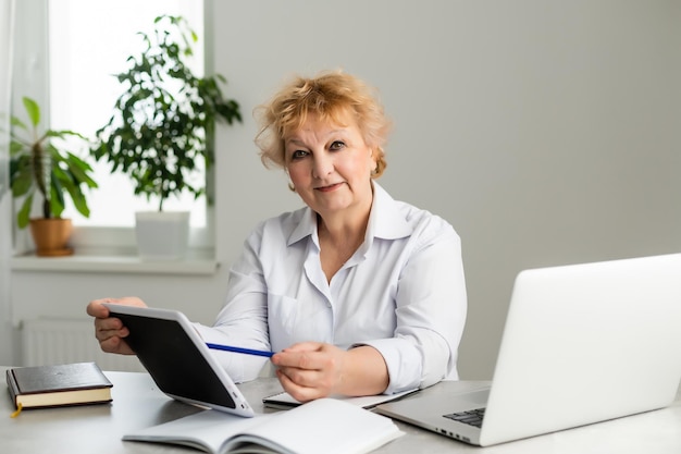 Senior vrouw thuis voor haar laptop die aantekeningen maakt tijdens het kijken naar een online Engelse taalles door een vrouwelijke leraar, e-learning concept.