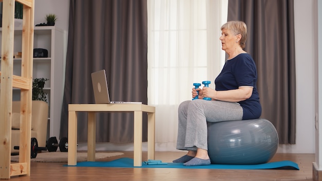 Senior vrouw training met gewichten zittend op stabiliteitsbal tijdens online fitnessles. Online training leertechnologie oude vrouw tillen training gezonde levensstijl sport fitness workout thuis