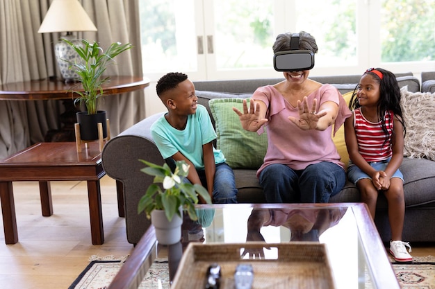 Senior vrouw van gemengd ras en haar kleinkinderen genieten samen van hun tijd thuis, zittend op een bank, met een VR-headset en glimlachend