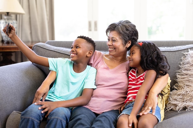 Senior vrouw van gemengd ras en haar kleinkinderen genieten van hun tijd thuis samen, zittend op een bank, omhelzen, glimlachen, een smartphone gebruiken, een selfie maken