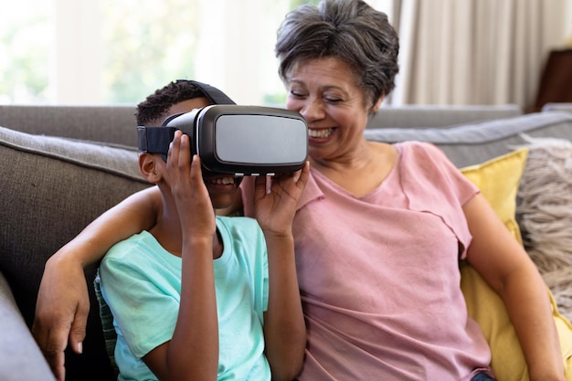 Senior vrouw van gemengd ras en haar kleinzoon genieten samen van hun tijd thuis, zittend op een bank, met behulp van een VR-headset, omhelzen en glimlachen