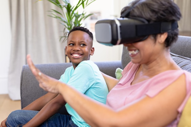 Senior vrouw van gemengd ras en haar kleinzoon genieten samen van hun tijd thuis, zittend op een bank, met behulp van een VR-headset, omhelzen en glimlachen