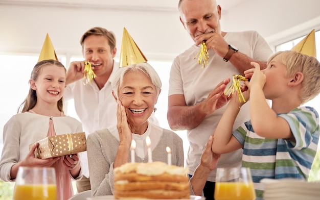 Senior vrouw viert haar verjaardag met familie thuis, draagt feestmutsen en blaast op fluitjes Oma kijkt naar verjaardagstaart en ziet er vrolijk uit terwijl ze wordt omringd door haar kleinkinderen en zoon