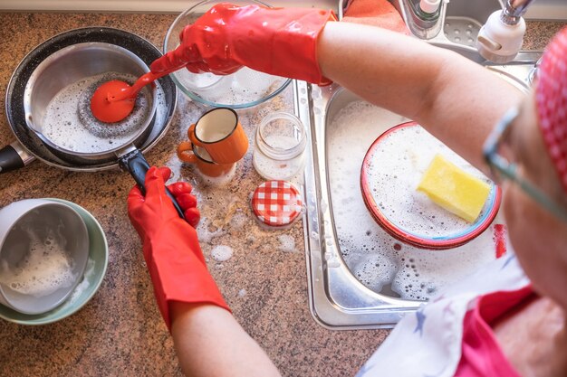 Senior vrouw wast servies voor het raam met rode handschoenen en bandana