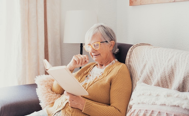 Senior vrouw zit binnenshuis in een gezellige bank met een open boek dat een spannend verhaal leest dat ontspannen is