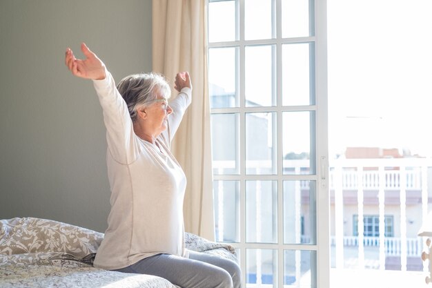 Senior vrouw zittend op bed met uitgestrekte armen en ogen gesloten in slaapkamer naast raam in haar appartement. tevreden oude vrouw die handen op bed thuis uitrekt