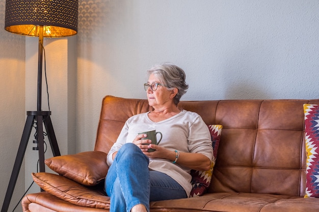 Senior vrouw zittend op de bank en koffiemok vast te houden terwijl ze thuis wegkijkt. Oudere vrouw ontspannen op de bank in de woonkamer. Doordachte vrouw die thuis koffie op de bank heeft