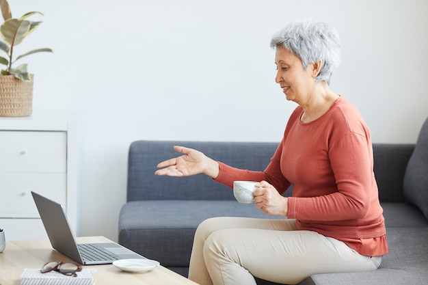 Senior vrouw zittend op de bank koffie drinken en heeft online conferentie op laptopcomputer thuis