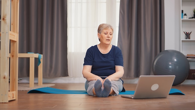 Senior vrouw zittend op yoga mat stretching boyd zittend op de mat. Online leren en studeren, actieve gezonde levensstijl sportieve oude persoon training workout thuis wellness en indoor training