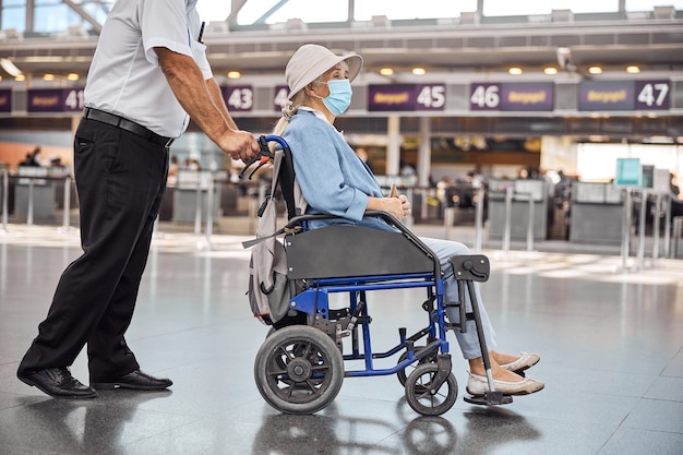 Senior vrouwelijke passagier met een gezichtsmasker en een hoed zittend in een transportstoel