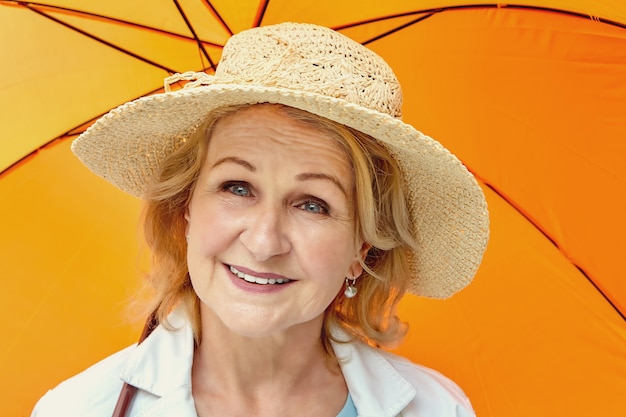 Senior white attractive blond woman about 62 years old in hat is standing under orange umbrella in the daytime.