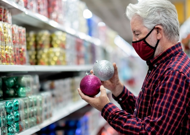 A senior white haired man holding two glittery christmas balls, silver and purple, wearing a medical mask due to coronavirus infection