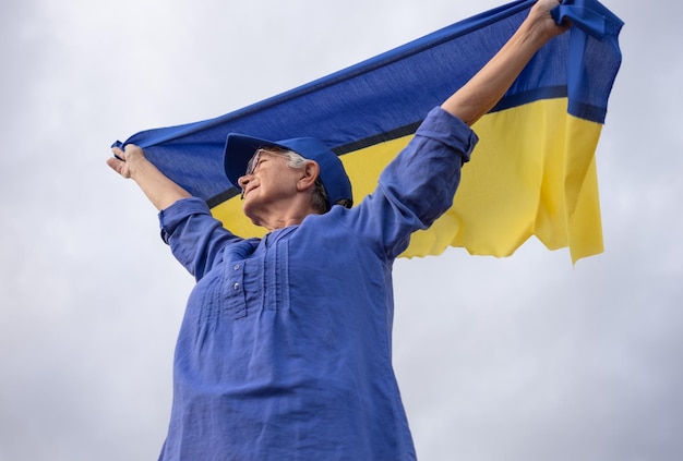 Senior whitehaired woman standing outdoors waving the Ukrainian flag looking away feeling the freedom No war stop fights we want peace