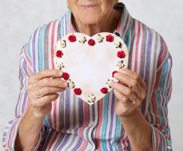 Senior woman between 70 and 80 years old keeps heart in the hands. Gray  background