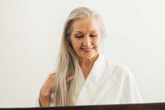 Senior woman in bathrobe looking down while adjusting her long white hair while in front of a mirror