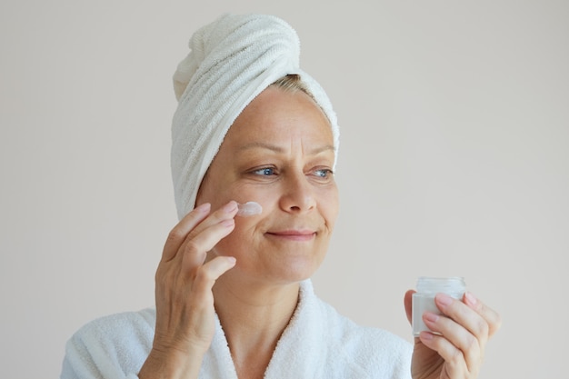 Senior woman in a bathrobe with a white towel on her head uses an antiaging face cream