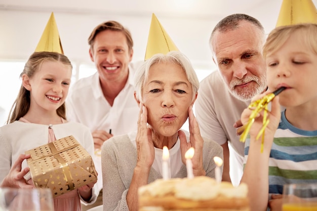 家族と一緒に誕生日を祝う年配の女性が、パーティーハットをかぶって笛を吹くおばあちゃんがバースデーケーキを見て、孫の夫と息子に囲まれて喜んでいる