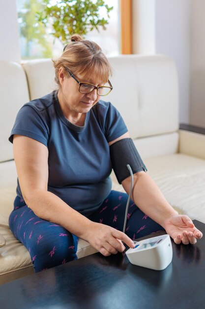 소파에 앉아 혈압계를 사용하여 고혈압으로 고통받는 노인 여성 집에서 혈압 수준을 확인하는 노인 여성