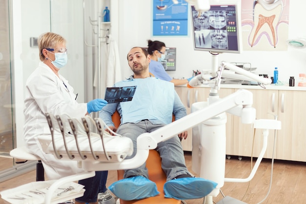 Senior woman dentist treating sick man patient in modern stomatological dentistry office showing teeth radiography explaining dental intervention