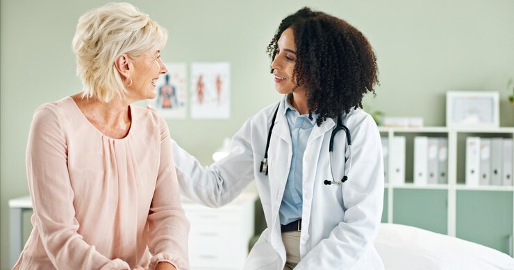 Photo senior woman doctor and comfort in consultation with discussion smile and good news at wellness clinic medic people and happy with support counselling and review for health on bed at hospital