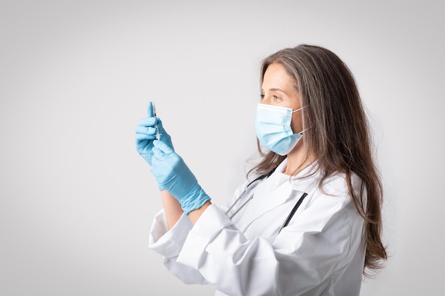 Senior woman doctor in white coat medical mask and protective gloves taking vaccine at syringe light