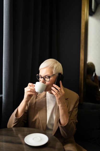 彼女の電話で話している間カフェでコーヒーを飲む年配の女性