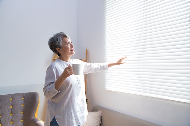 Senior woman drinking coffee next to the window.