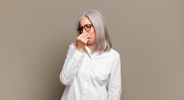 Senior woman feeling disgusted, holding nose to avoid smelling a foul and unpleasant stench