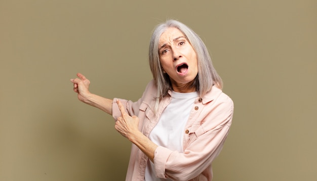 Senior woman feeling shocked and surprised, pointing to copy space on the side with amazed, open-mouthed look